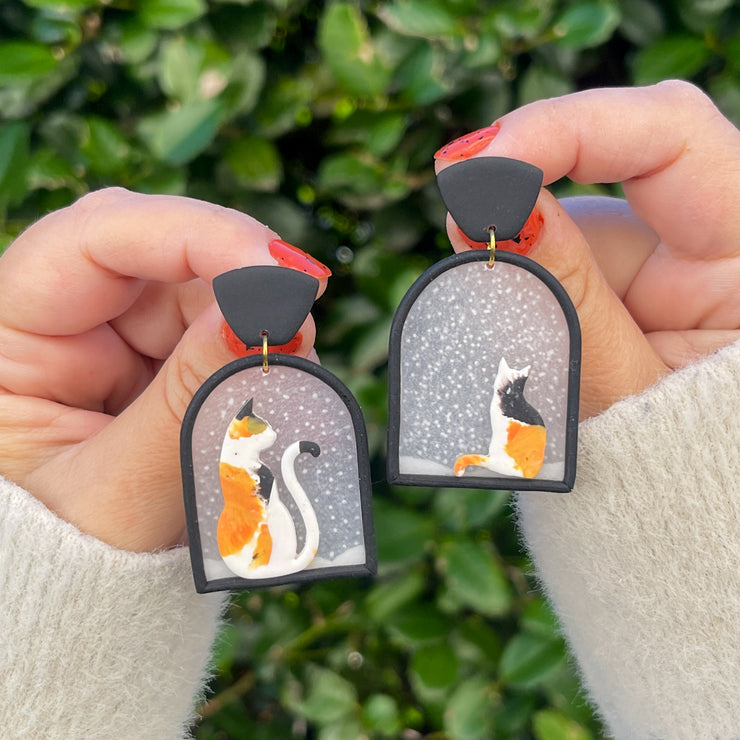 Polymer clay earrings design of two calico cats seating in a window watching snow fall 